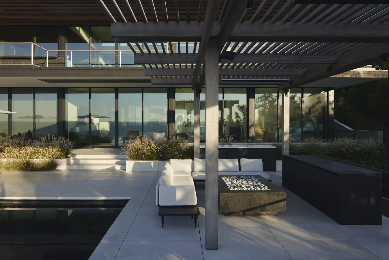 Outdoor seating area with a sectional sofa and fire pit under a pergola beside a reflecting pool at Ocean Bluff by Stelle Architects.