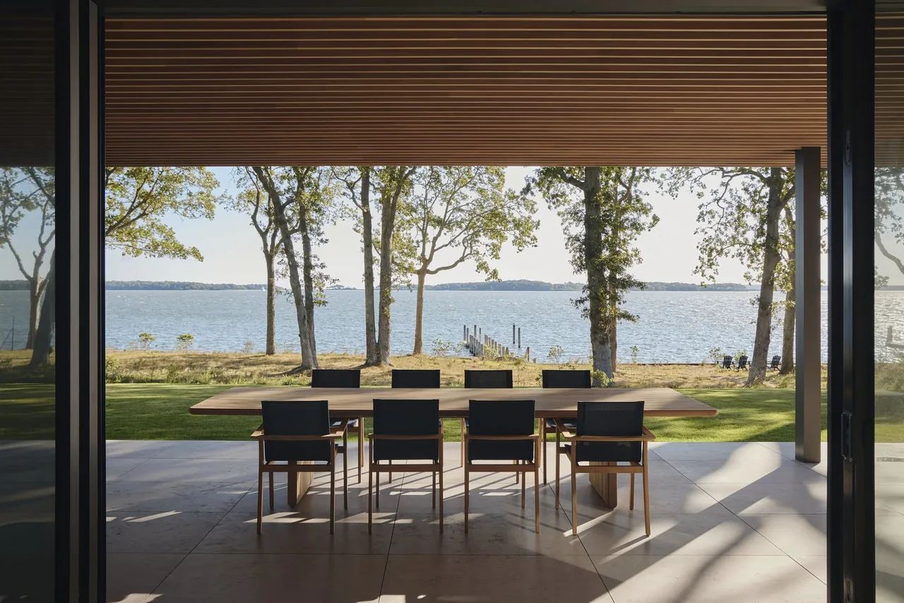 Outdoor dining space under a horizontal wood slat ceiling with views of a lake and dock at Forest Edge project.