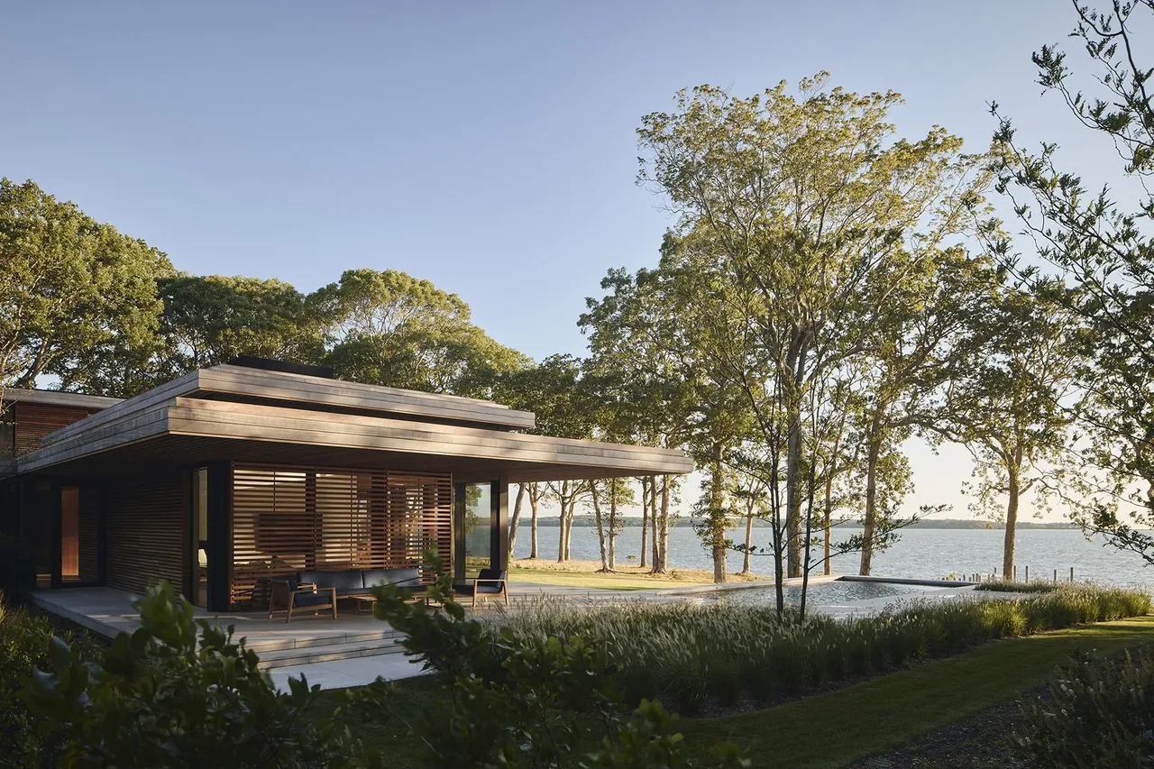 The FOREST EDGE project features a contemporary house with horizontal wood slats and a flat roof overlooking a reflective pool and lake.