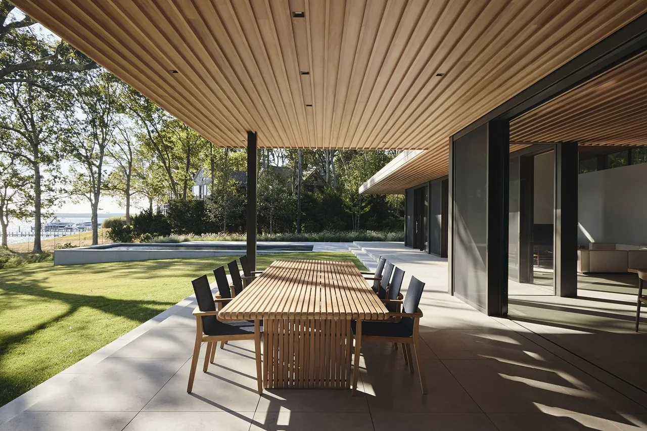 Covered patio dining area features a wooden slat ceiling and table with modern chairs overlooking landscaped lawn and water.
