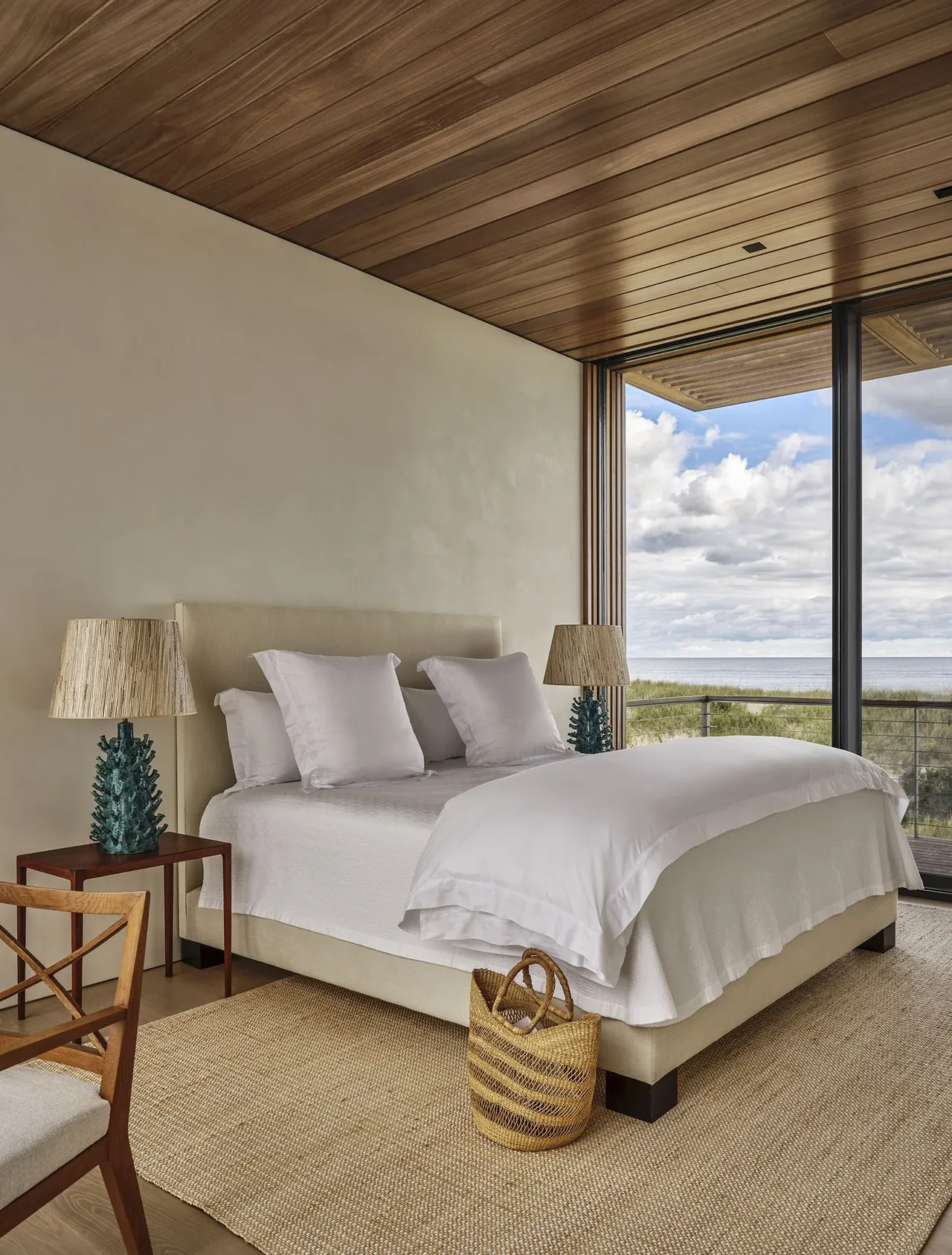 Bedroom with floor-to-ceiling window overlooking ocean, featuring wood-paneled ceiling and a bed with white linens.