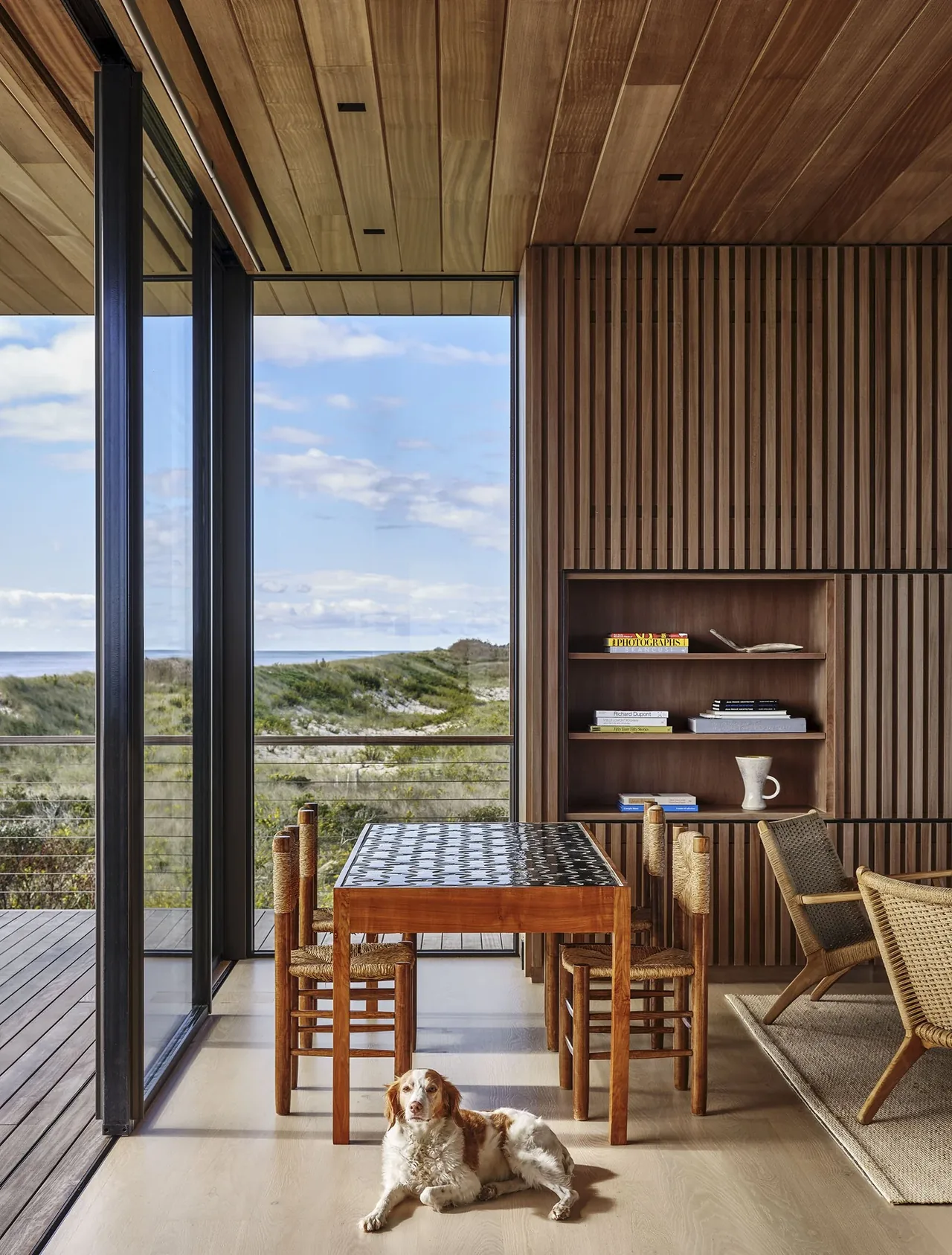 Seaside project dining space features wood paneling, large glass walls with ocean view, and woven seating by Stelle Architects.