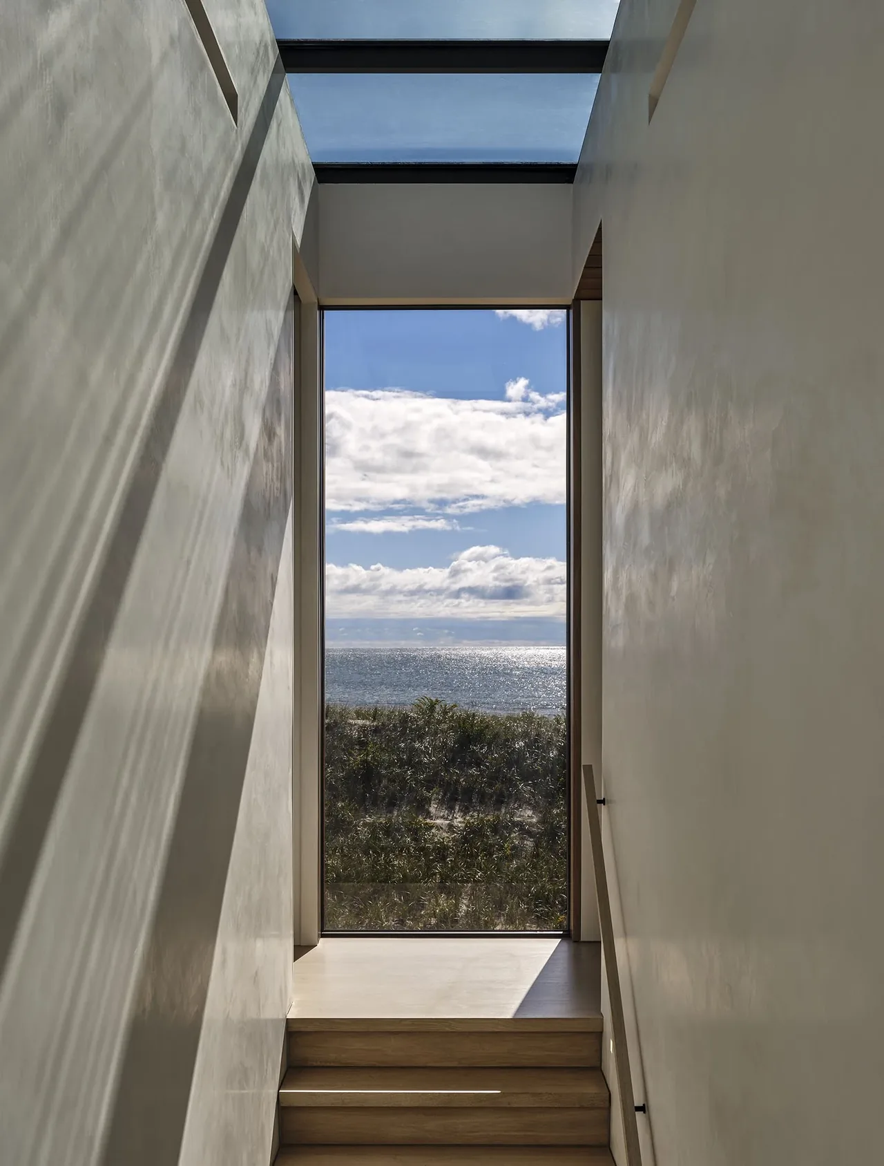 A narrow stairway features a large floor-to-ceiling window framing an ocean view under a partly cloudy sky.