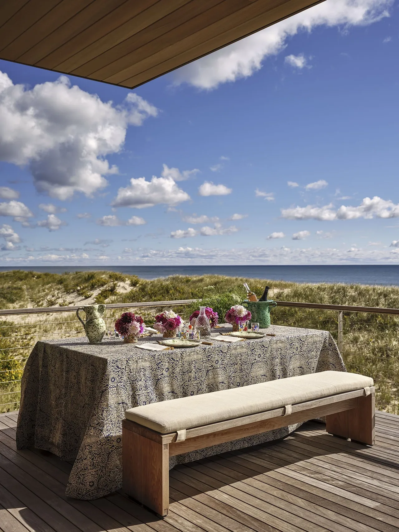 Seaside project outdoor dining space features a wooden bench and patterned tablecloth with an ocean view.
