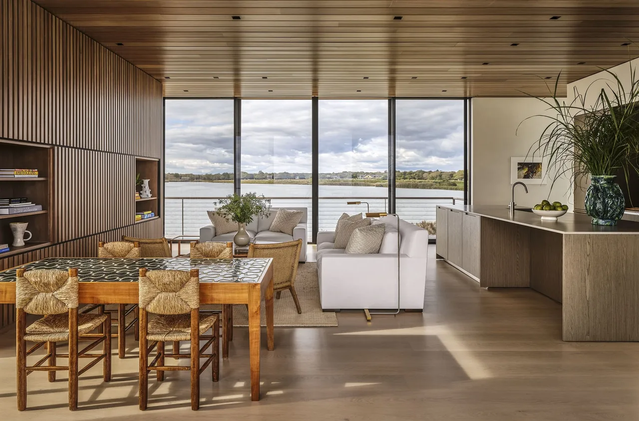 Seaside project features an open plan kitchen and living area with wood cladding and large windows framing ocean views.