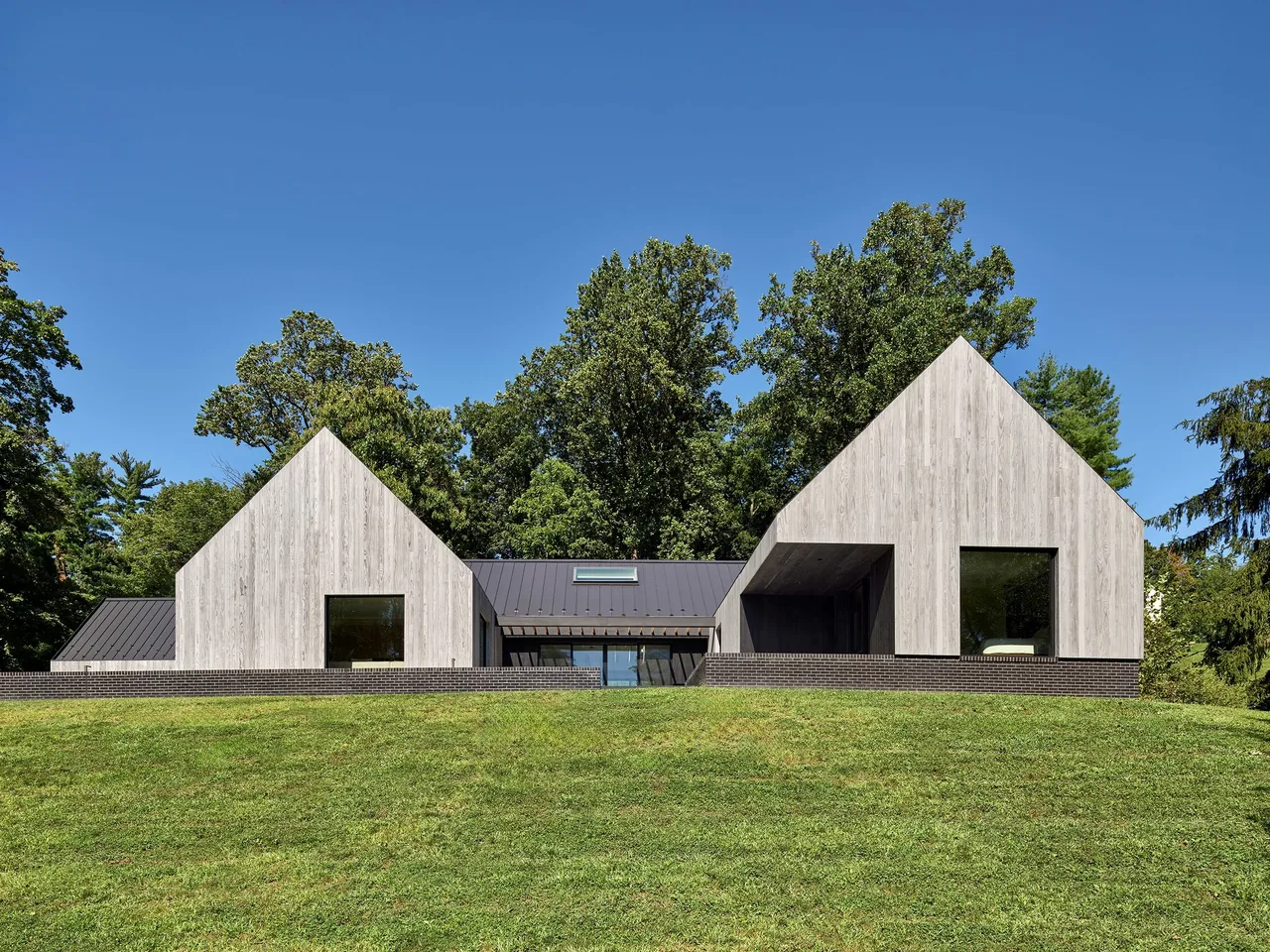 Gladwyne Residence by Stephen Varenhorst Architects features connected volumes with wood cladding and large windows on a grassy slope.