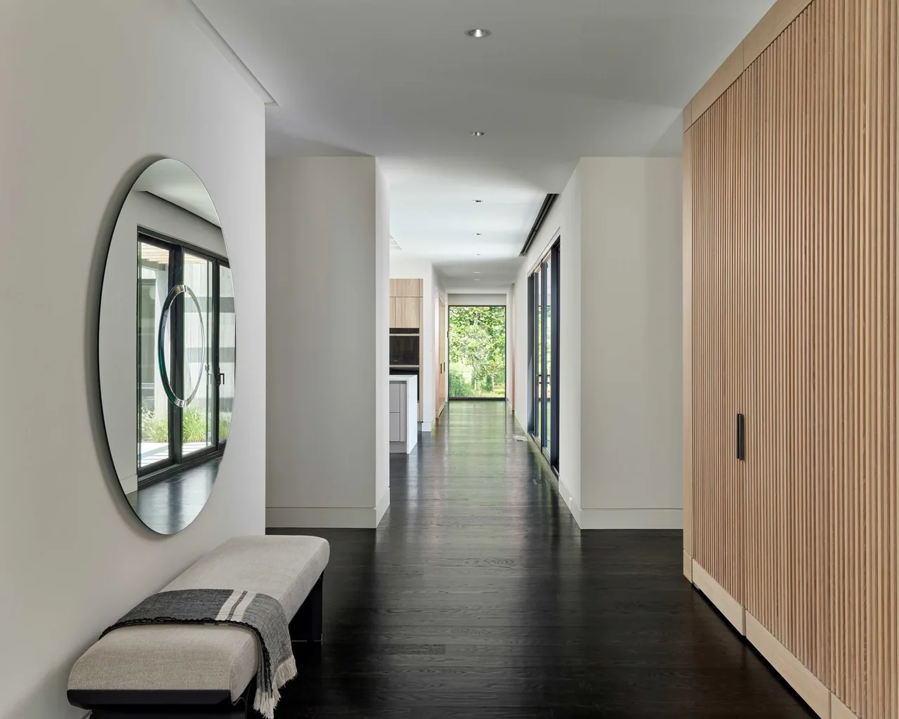 Hallway space in the Gladwyne Residence features dark wood flooring, slatted wood closets, and a large round mirror.