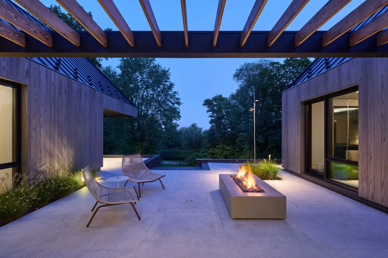 Patio space with a fire pit, wooden pergola, and seating between two parts of the residence at dusk.