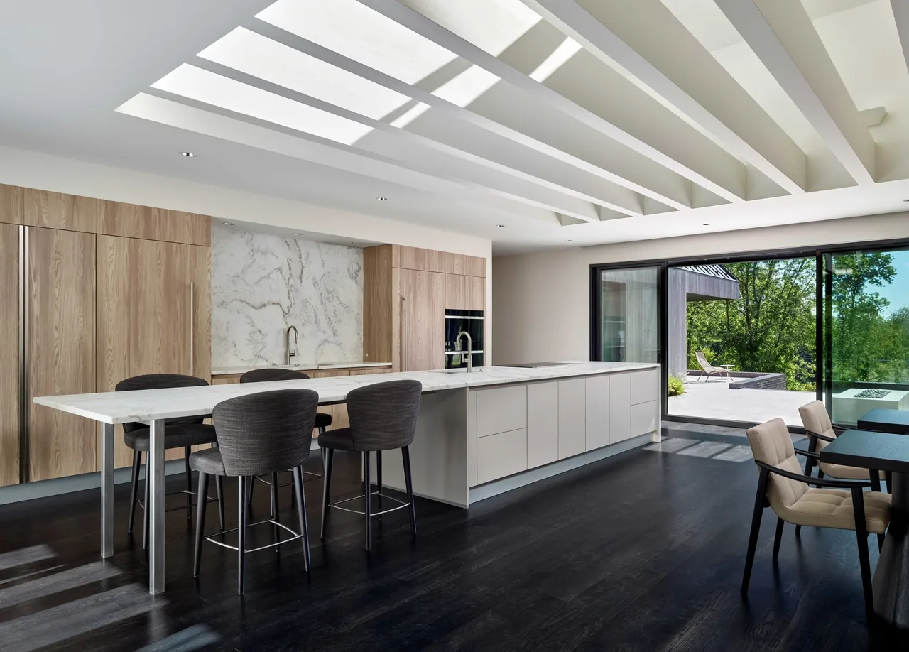 The Gladwyne Residence kitchen features a large marble island, wood-clad cabinetry, and expansive sliding glass doors to an outdoor seating area.