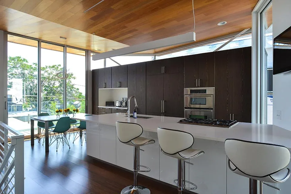 Open kitchen with a white island, dark wood cabinets, wood-paneled ceiling, and floor-to-ceiling windows.