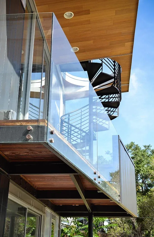 Modern balcony features a glass railing and black metal spiral staircase beneath a wood soffit.