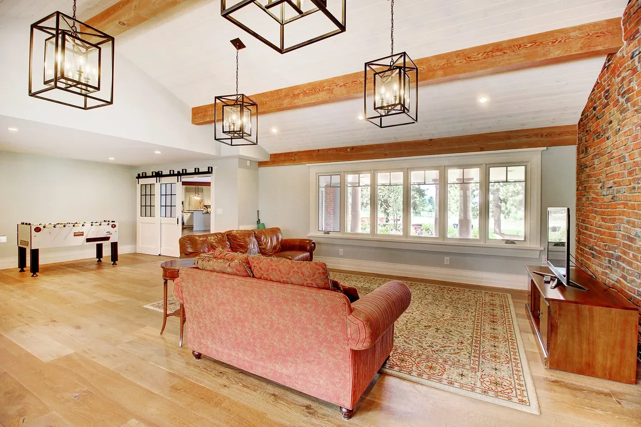 Living room featuring vaulted ceiling with wood beams, large multi-pane windows, brick accent wall, and traditional furniture.
