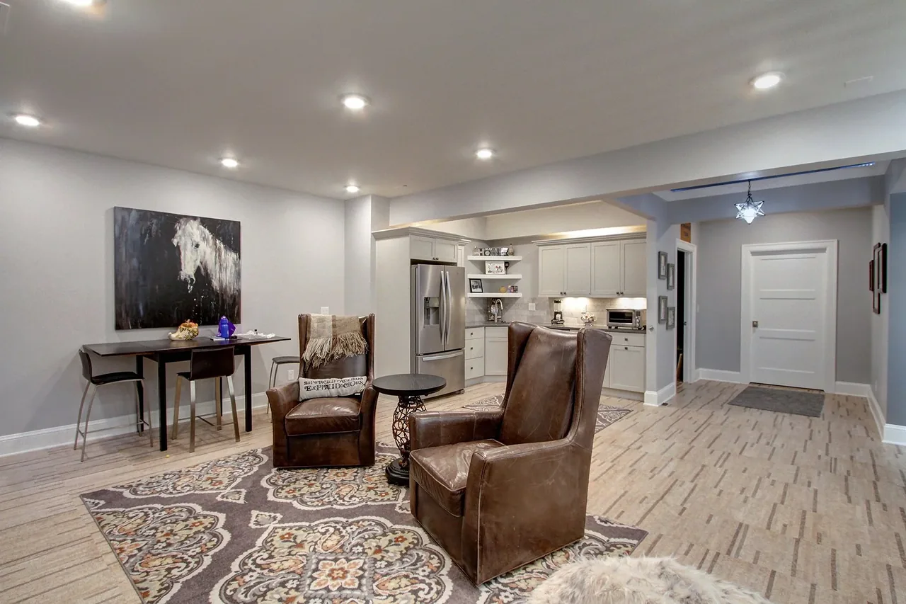 This open layout connects a living area with leather chairs to a kitchen featuring gray cabinets and stainless steel appliances.