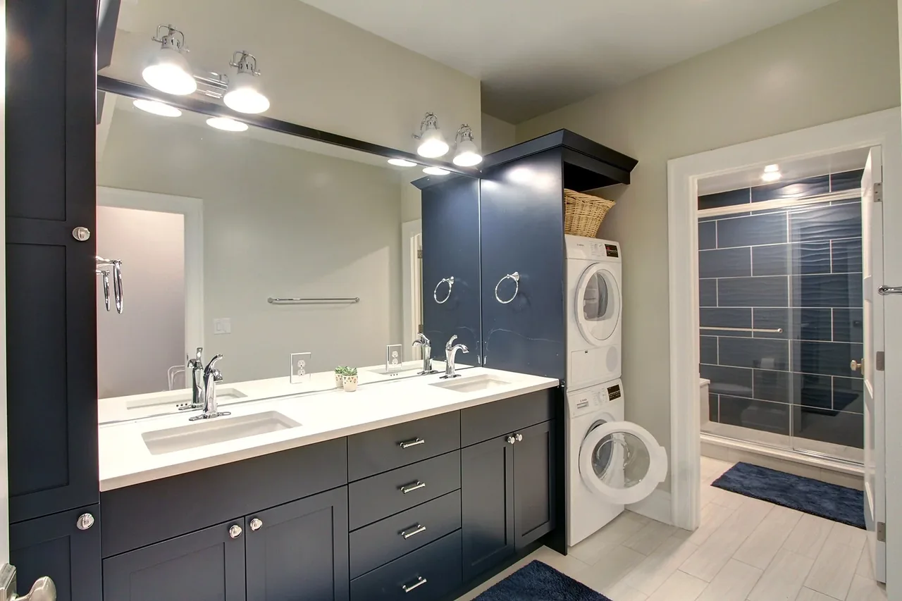 Bathroom includes dark blue cabinets, double sinks, and a stacked washer and dryer near a shower with glass doors.