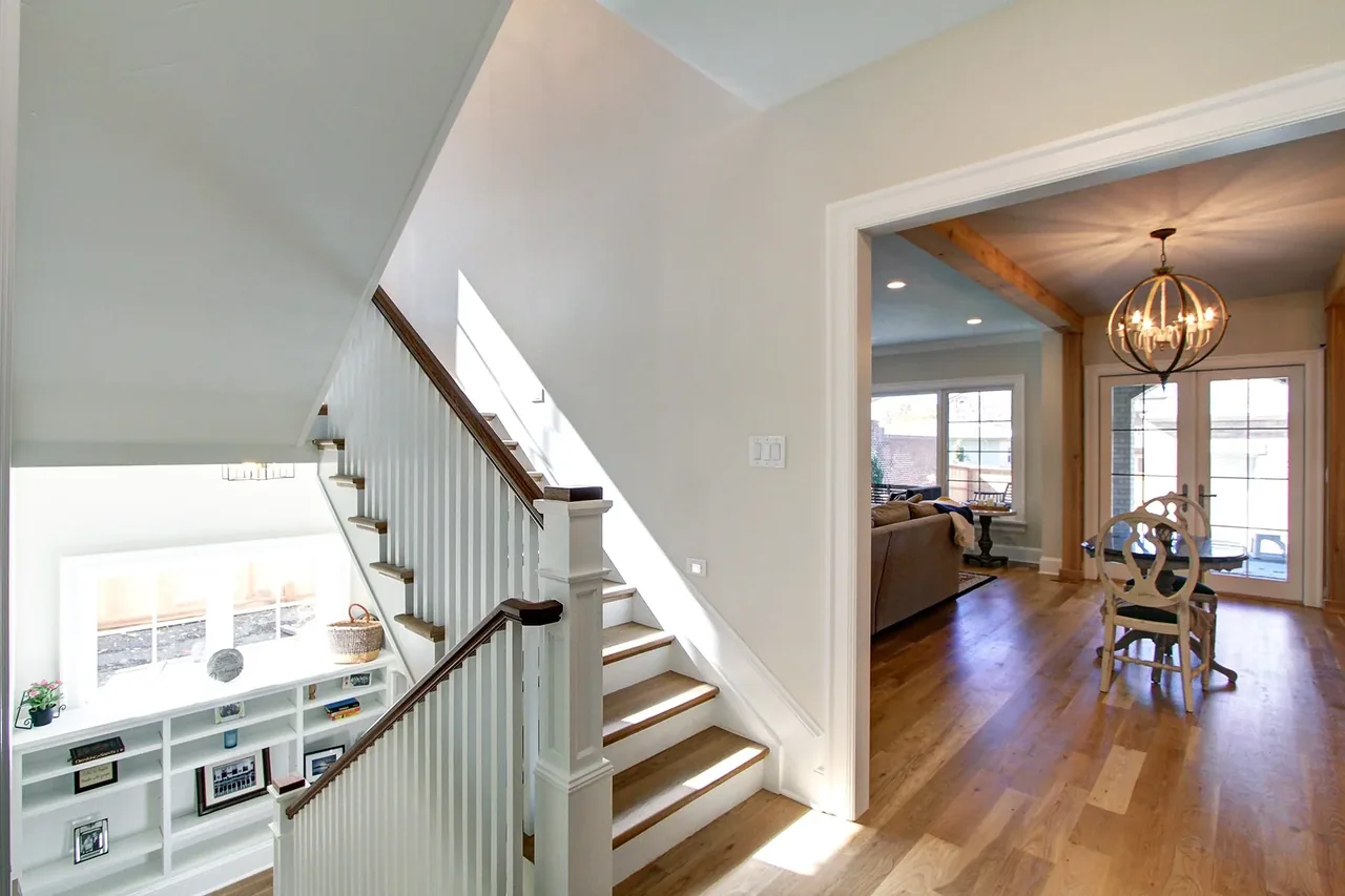 A white staircase with wood treads leads from a hallway into an adjacent dining and living area with hardwood floors and a spherical chandelier.