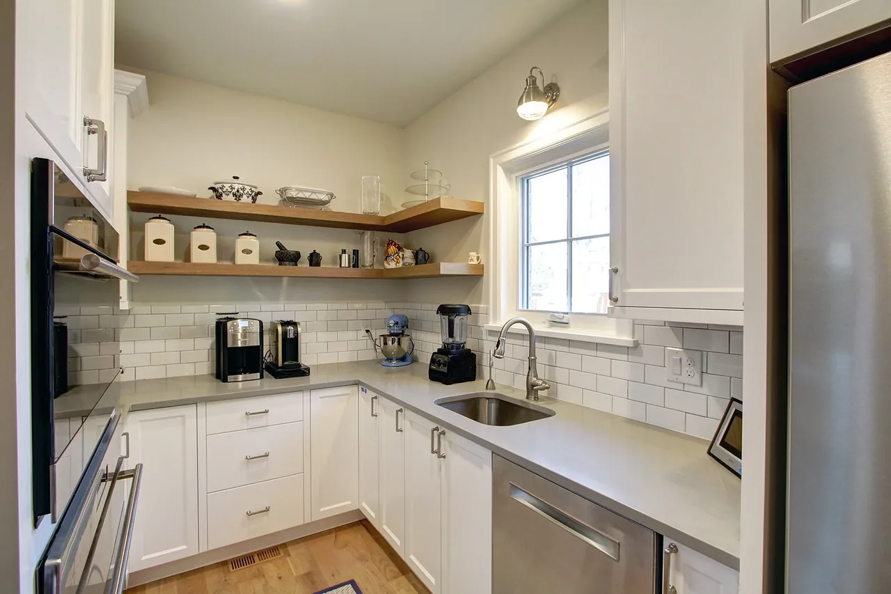 Compact kitchen featuring white cabinets, subway tile backsplash, wood shelves, and stainless steel appliances below a window.