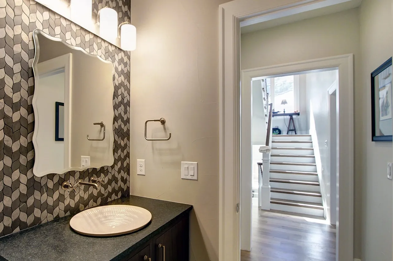 Bathroom vanity features patterned tile backsplash and a scalloped-edge mirror with an adjacent view of a staircase.