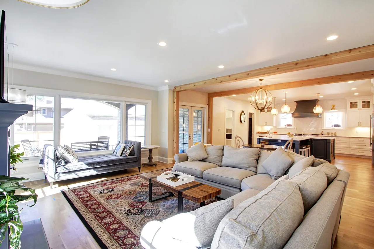 Living area with gray sectional sofa and wooden beams opening to kitchen with multiple pendant lights.