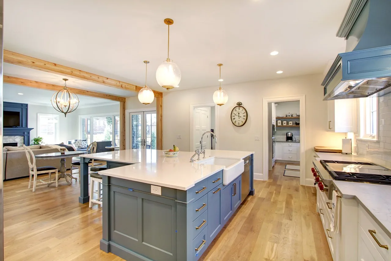 Kitchen with blue cabinetry island and white countertops opens to living and dining spaces with exposed wooden beams.