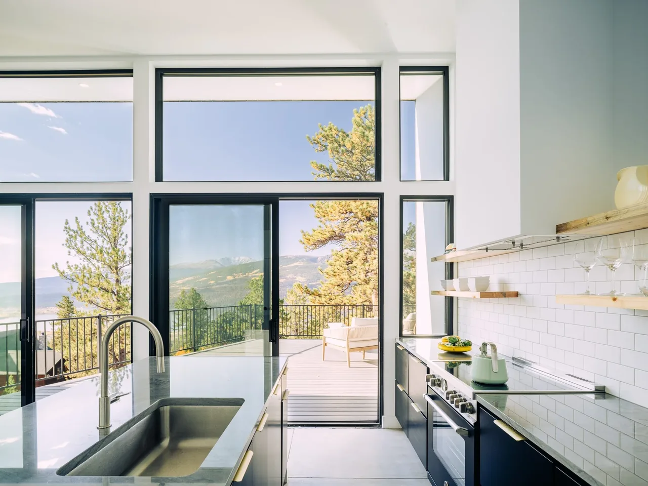 The kitchen features black cabinetry, open wood shelving, and floor-to-ceiling windows leading to an elevated deck with expansive mountain views.
