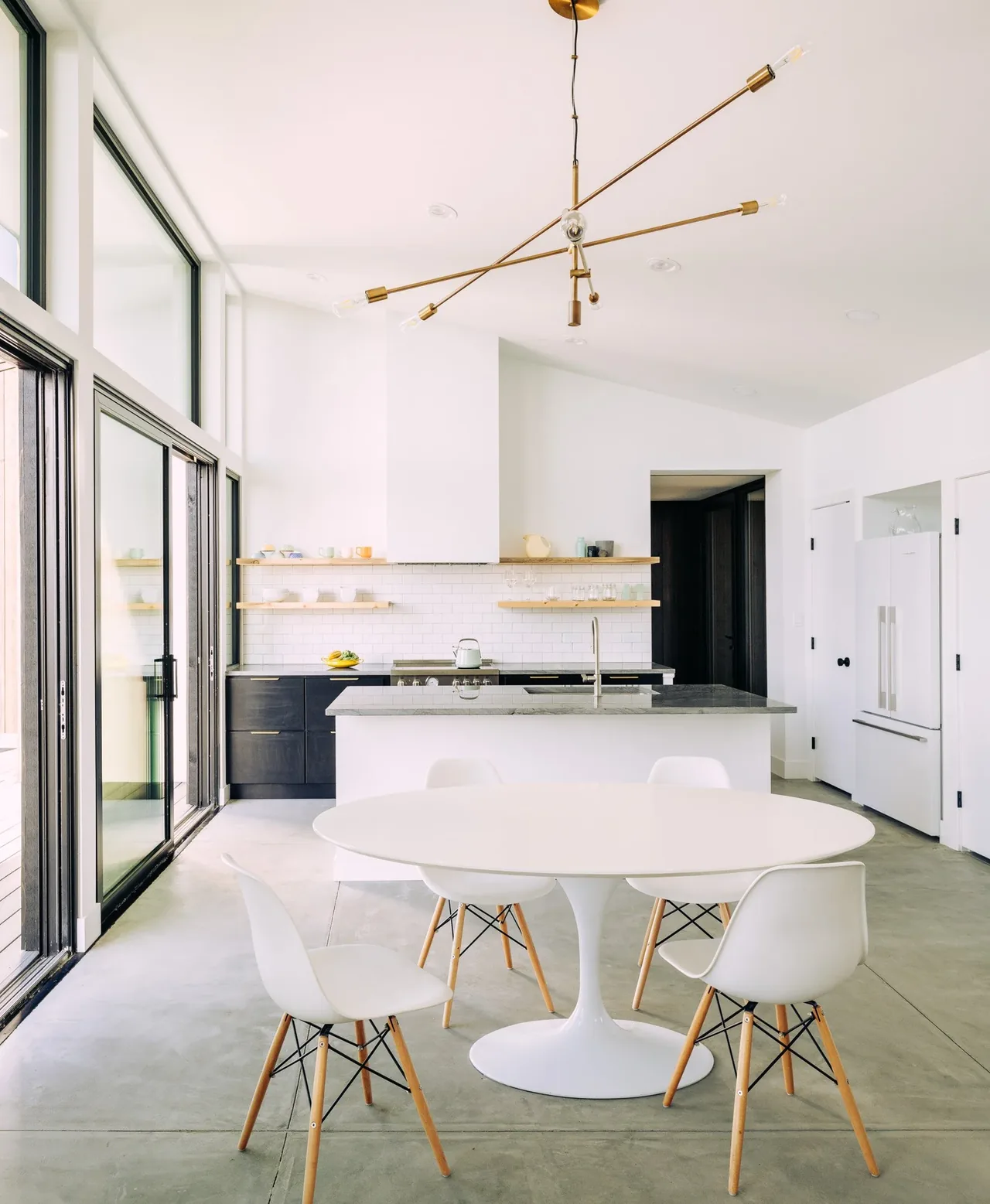 The kitchen and dining space features black lower cabinets, open wood shelving, and a white round table with modern chairs.