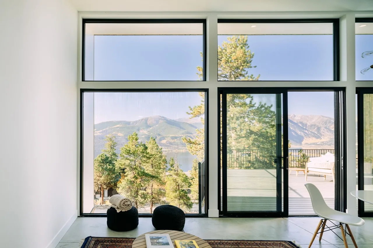 Room with floor-to-ceiling black-framed windows and sliding door opening to an elevated deck overlooking mountains and lake.