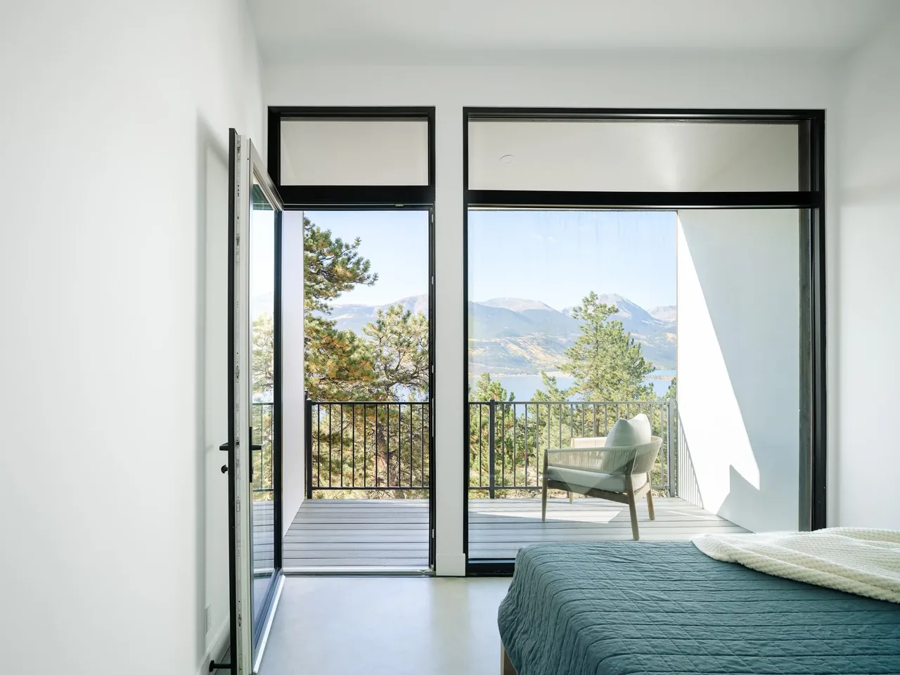 Bedroom with floor-to-ceiling black-framed windows and door opening to an elevated balcony overlooking mountains and a lake.