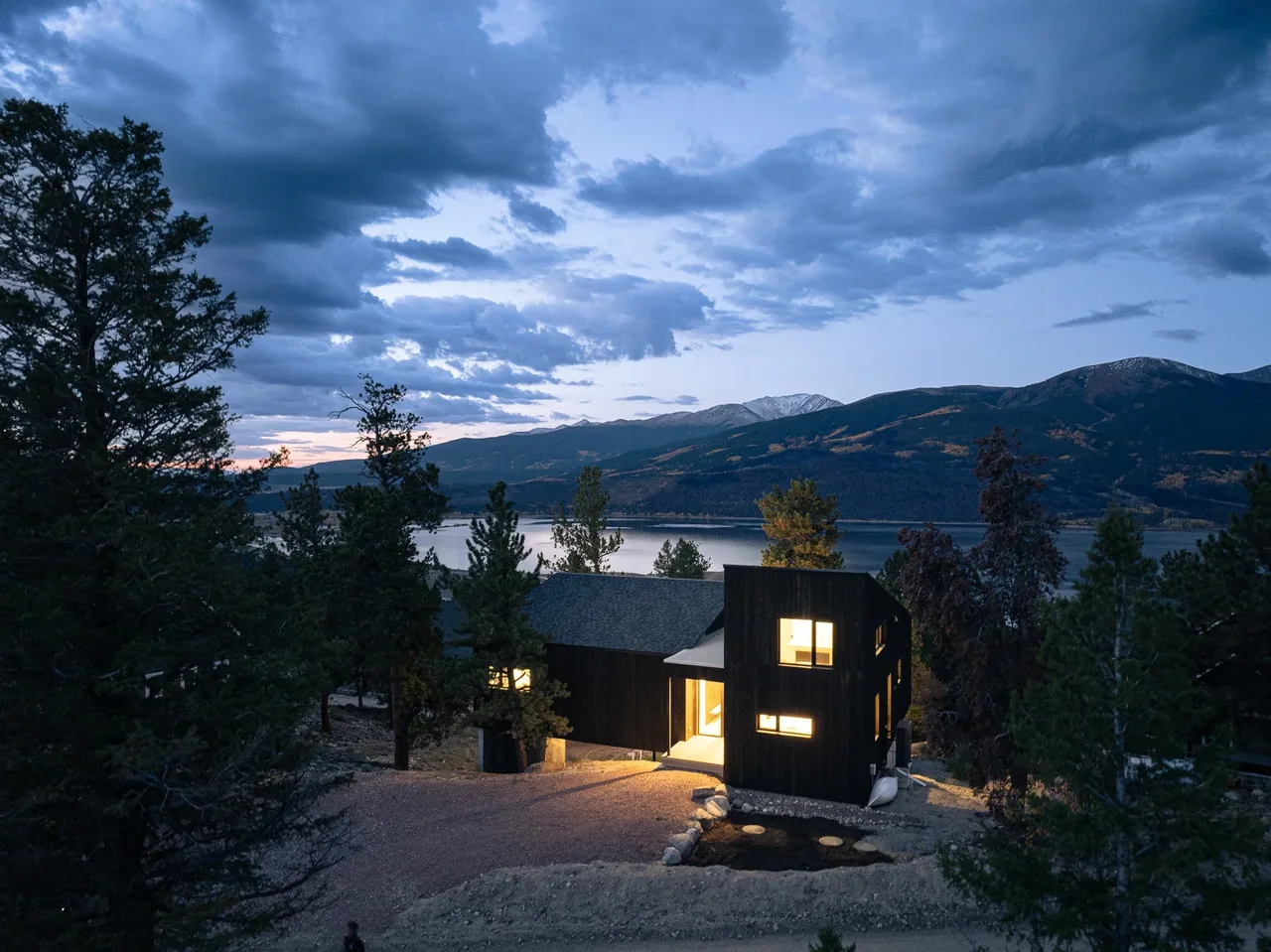 Twin Lakes Bothy features a dark wood exterior with multiple illuminated windows overlooking a lake and mountain landscape at dusk.