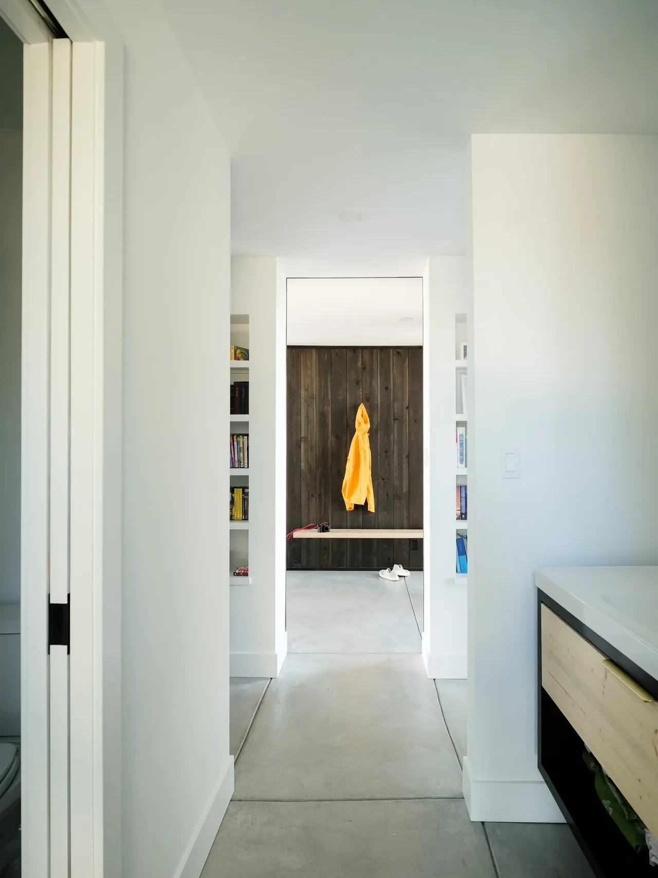 Hallway with concrete flooring and white walls framed by built-in shelving and a dark wood-paneled accent wall featuring a floating bench.