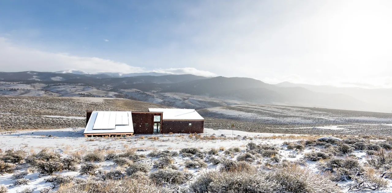Blue J is a house by Shape Architecture Studio with rust-colored siding and an overhanging roof, located on a snowy hilltop.