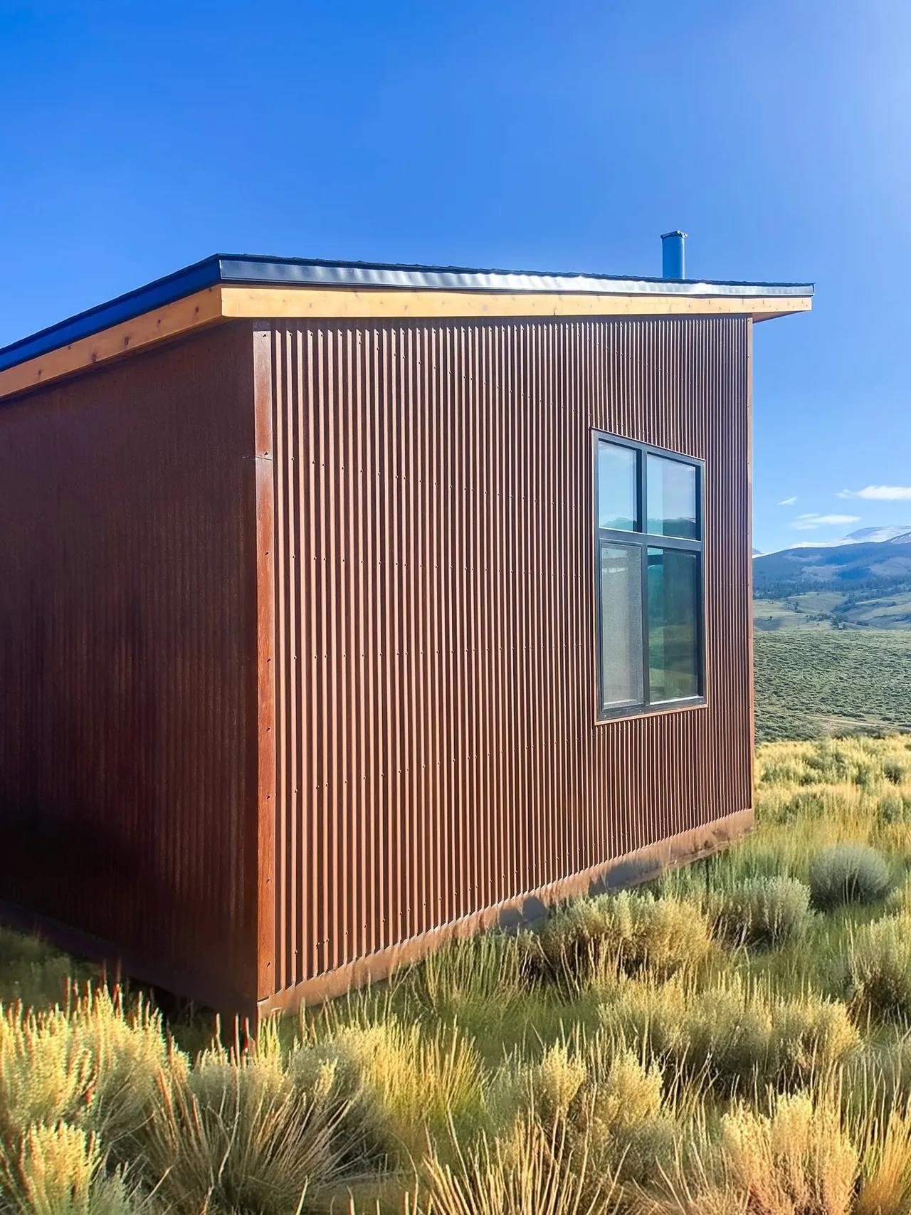 The Blue J project features rust-colored corrugated metal siding with an overhanging roof situated in a natural open landscape.