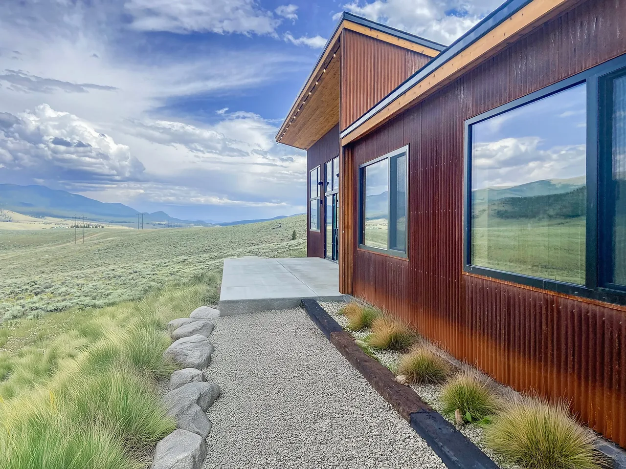 Blue J project features rust-colored corrugated metal siding with large black-framed windows overlooking open grassland and mountains.