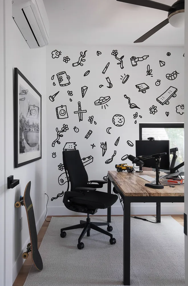 Home office with a wooden top desk, black office chair, skateboard, and wall adorned with whimsical black line drawings.