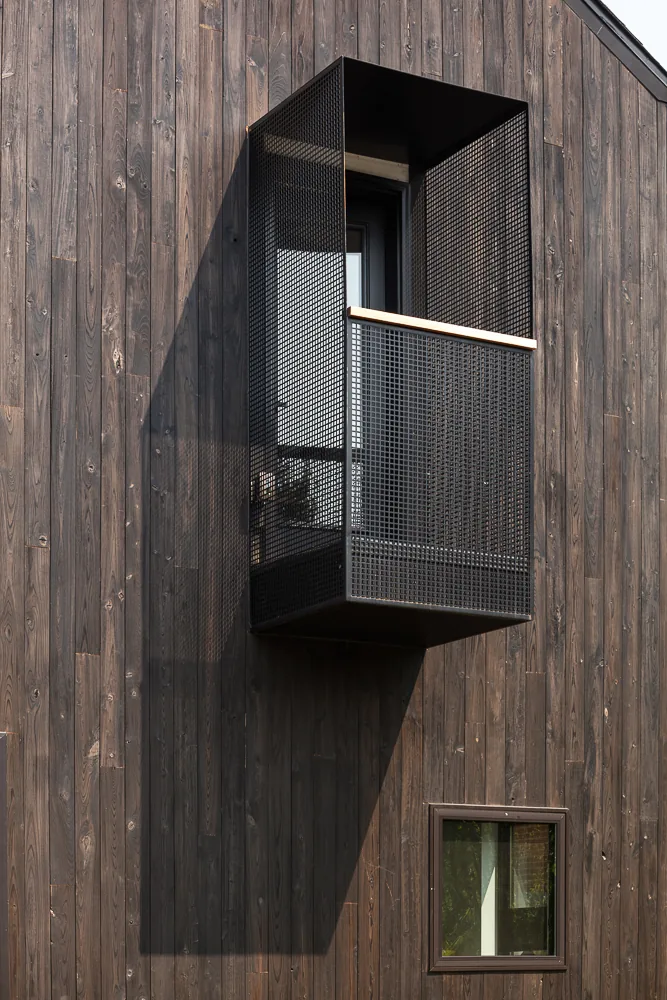 A projecting black metal mesh balcony contrasts with the dark vertical wood siding and a small square window on the exterior facade.