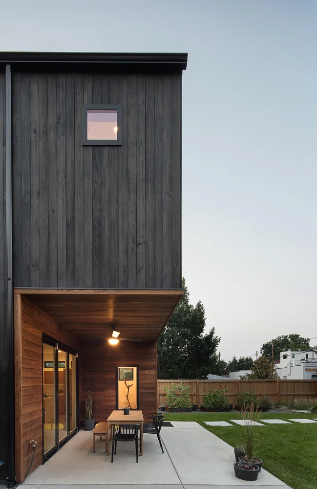 Two-story house with vertical dark siding above and horizontal wood siding below at the covered back patio with dining furniture.