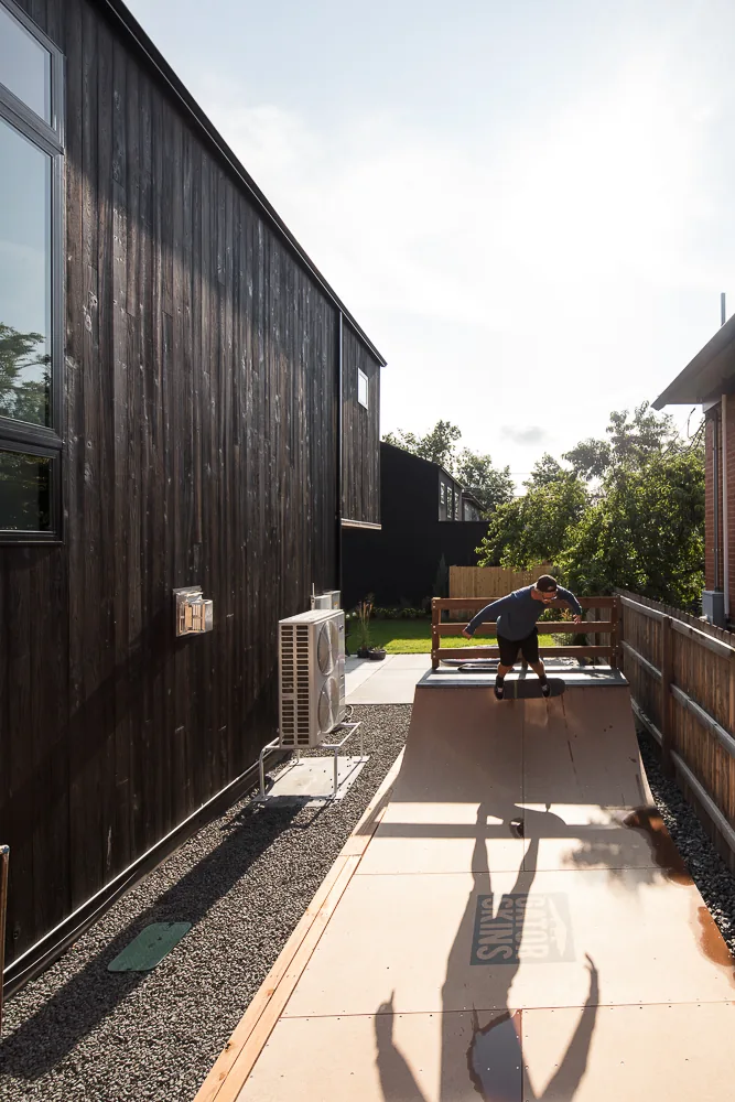 Side yard skate ramp made of wood panels next to a house clad in black wood siding with visible air conditioning units.