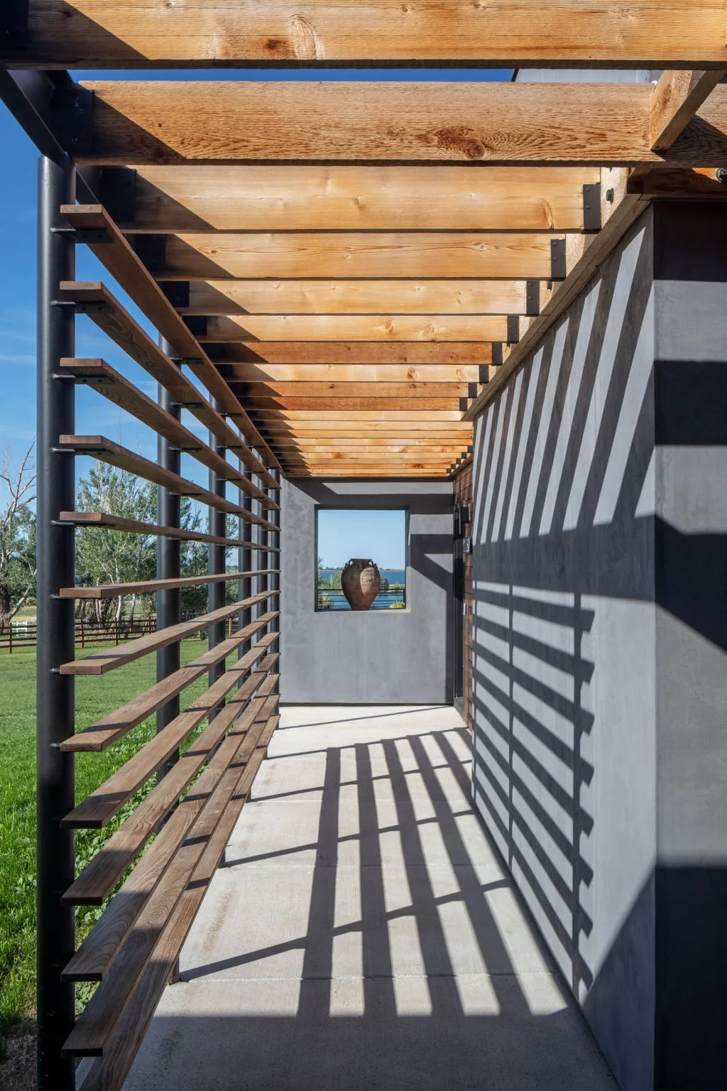 Outdoor corridor with wood slats and metal structure casting linear shadow patterns on a concrete floor and grey plastered wall.