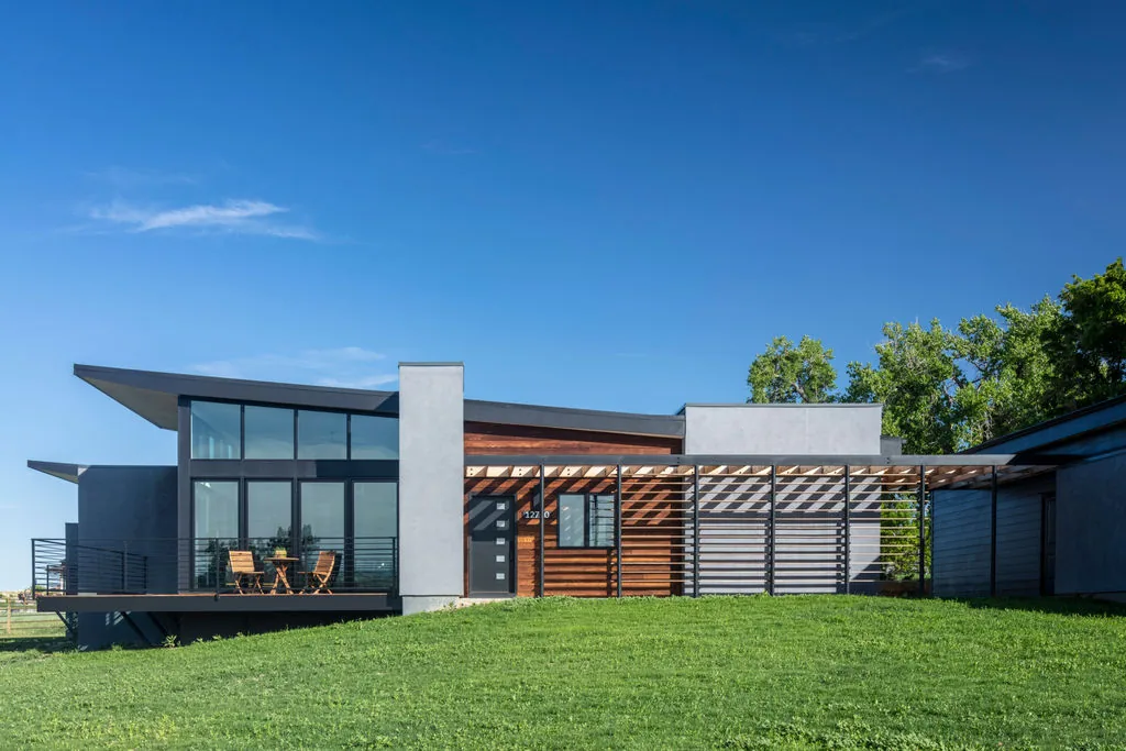 Hoff Residence features a modern design with large windows, metal railings, and a pergola creating striped shadows on the textured wall.