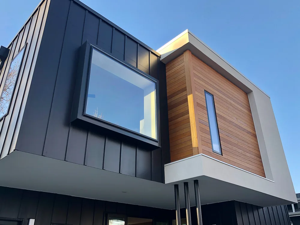 Dempster Residence features black metal panel siding and a cantilevered wood siding volume with vertical window.
