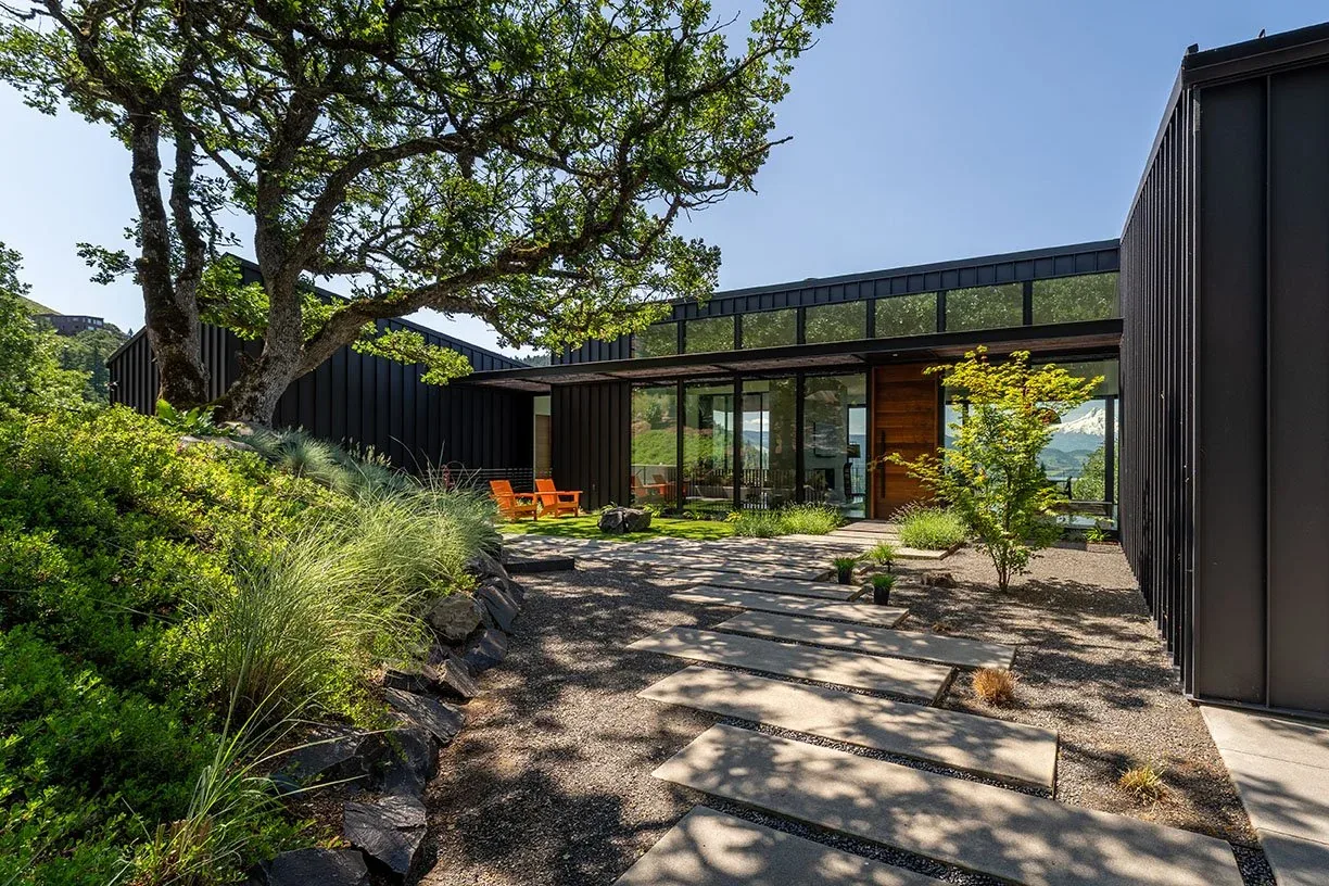Hidden Oaks Residence features black standing seam metal siding and floor-to-ceiling glazing in a hillside setting.