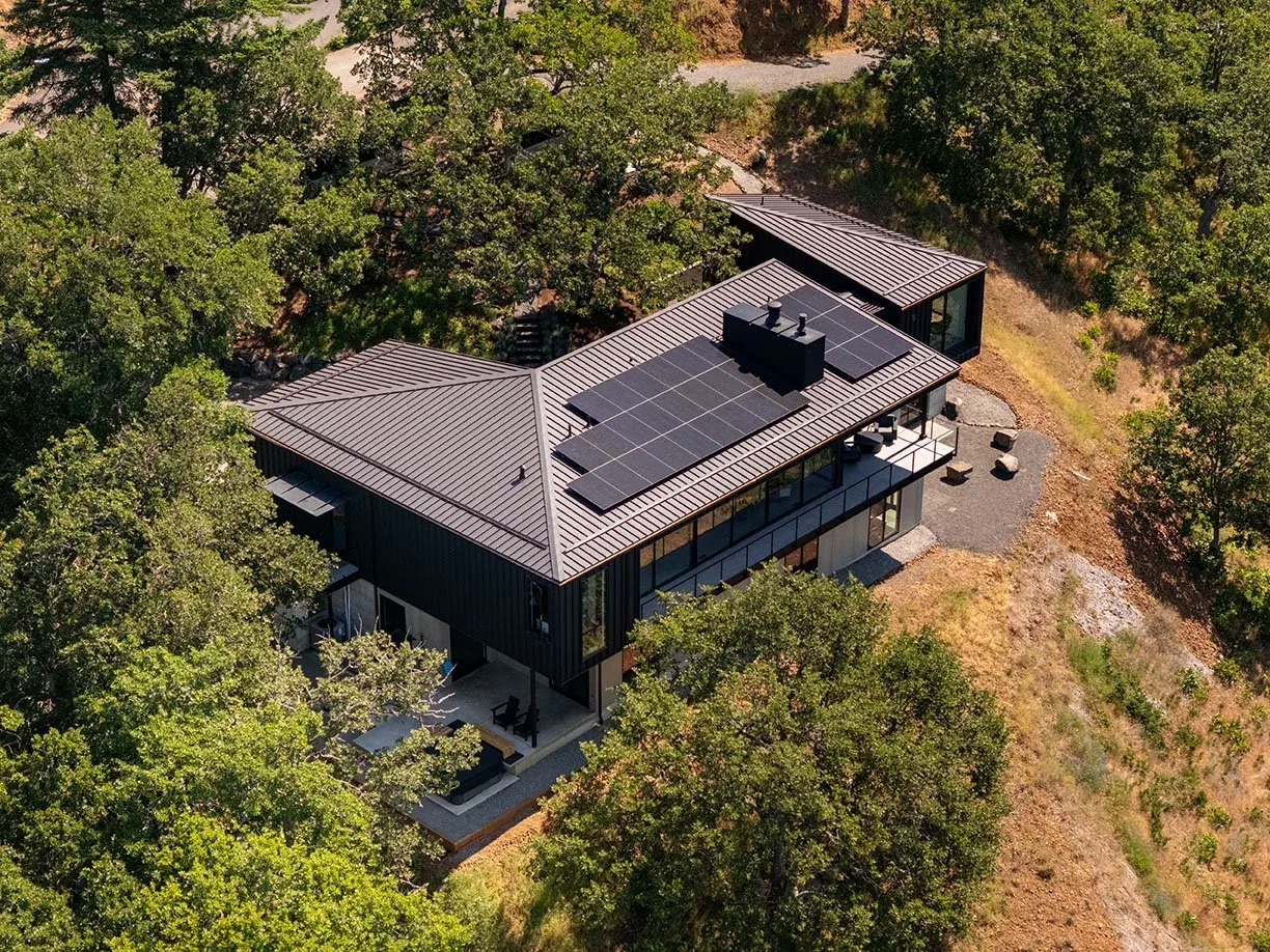 Hidden Oaks Residence features a black metal roof with solar panels, nestled in a wooded hillside landscape.