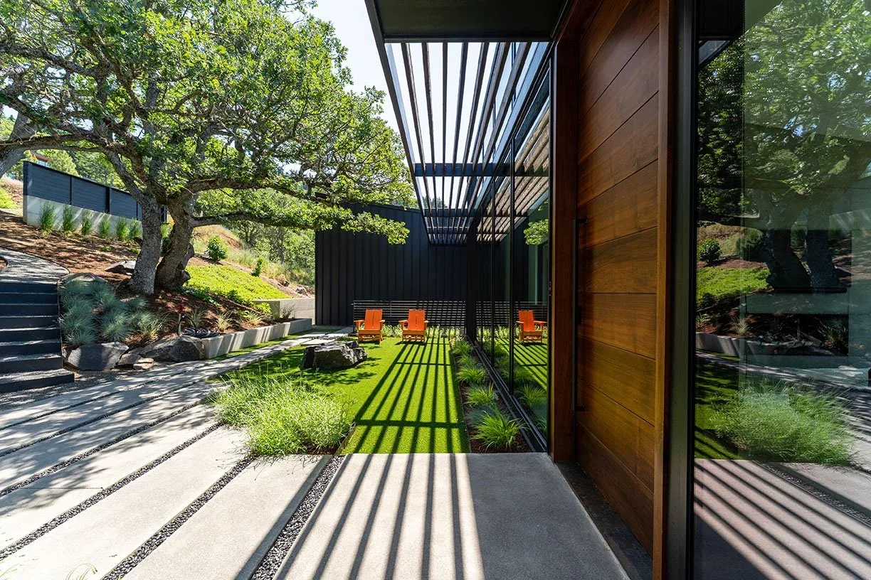 The Hidden Oaks Residence features an outdoor seating area on artificial turf with orange Adirondack chairs and mature oak trees, framed by black cladding and a slatted pergola.