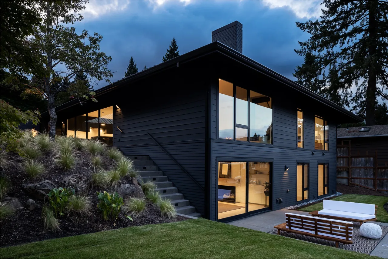 The Cardinal House features dark horizontal wood siding, large windows, and an outdoor seating area on a concrete patio at dusk.
