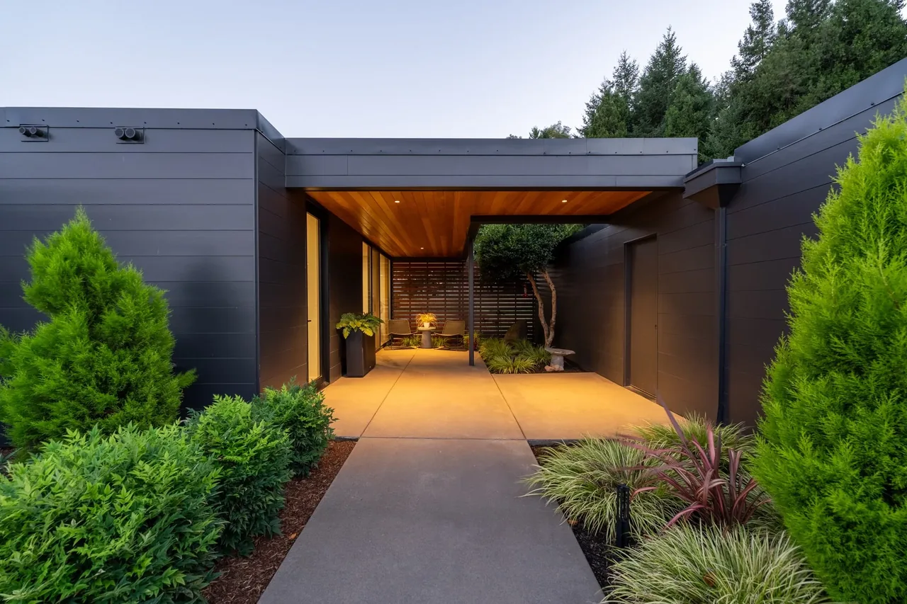 Modern single-story house entrance with black siding, covered walkway, and landscaped garden outside.