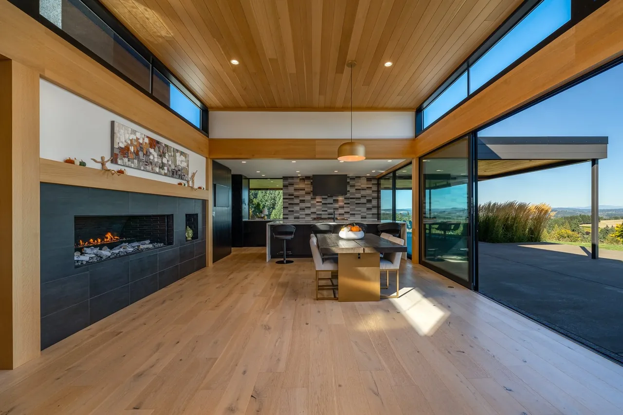 Burnt Tree Farm dining space features light wood flooring, a long dining table, and a fireplace with black tiles and black window frames.