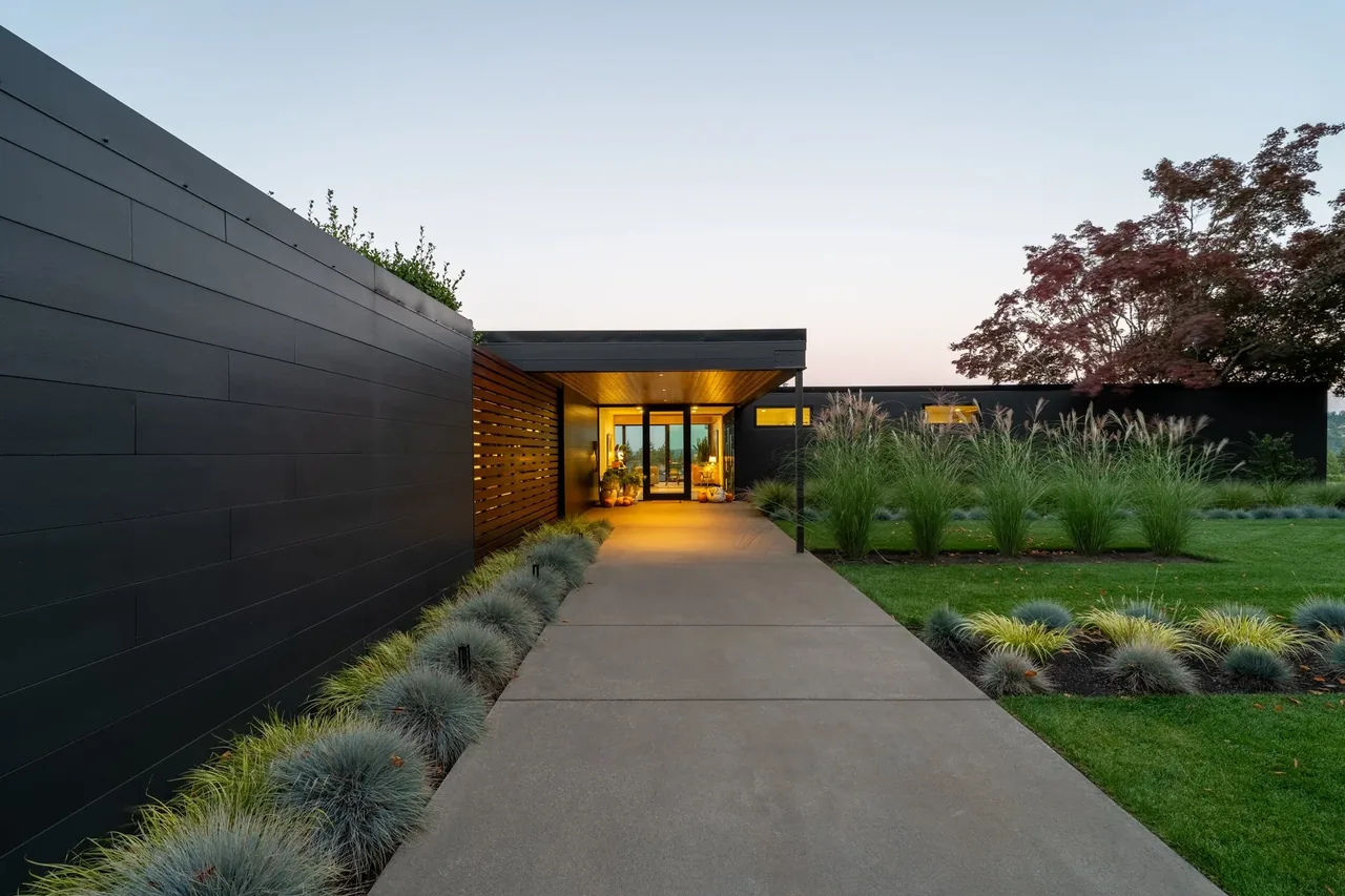 Entrance path to a modern single-story house at sunset, featuring black siding and landscaped greenery.