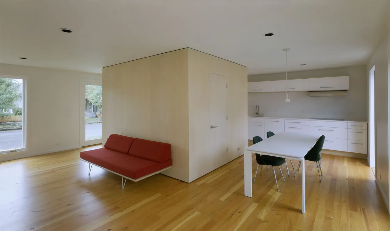 Open living and kitchen area with a central wood-paneled storage cube and minimalist furnishings in the Cape Cod project.