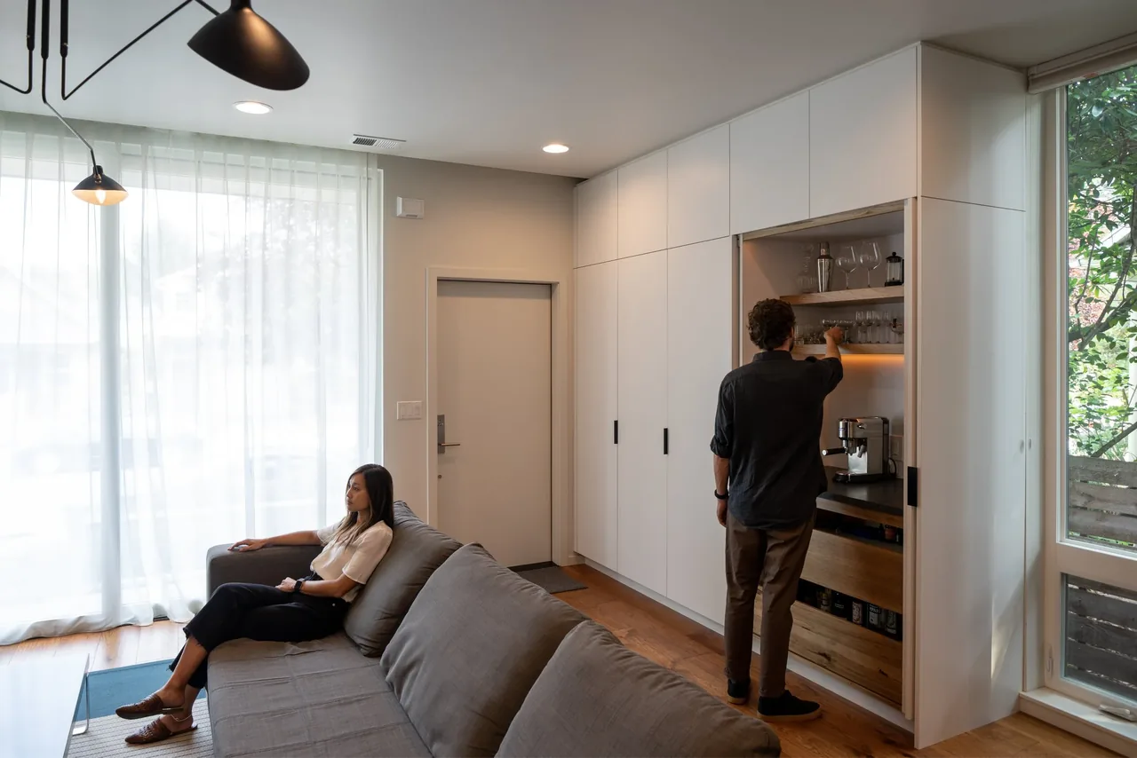 Living room with built-in white cabinetry coffee station and gray sofa, featuring wood flooring and large window with sheer curtains