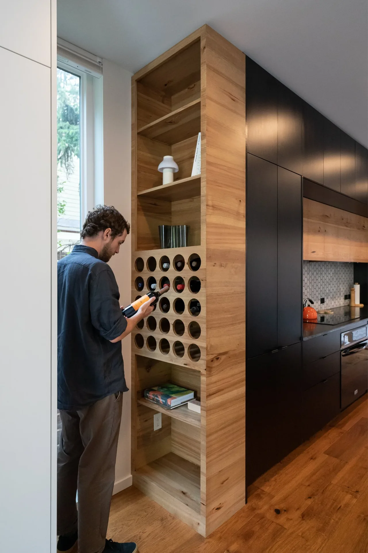 A man selects a wine bottle from a vertical wooden wine storage adjacent to black kitchen cabinetry with a hexagonal tile backsplash.