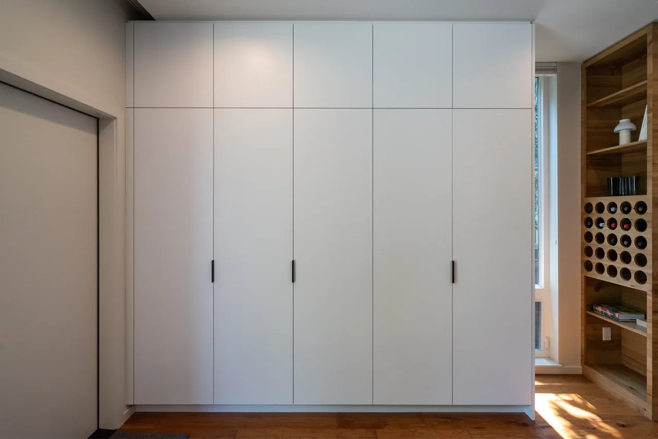 Built-in white storage cabinets with vertical handles, wood open shelving, and wine bottle storage in a modern interior space at Cleveland House.